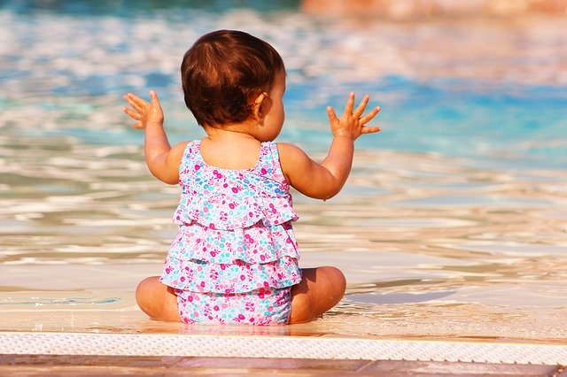 Baby sitting on the beach with hands up and fingers splayed. 